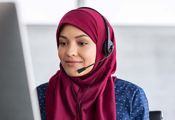 Yngre kvinde med tørklæde og headset arbejder på computer i callcenter. 